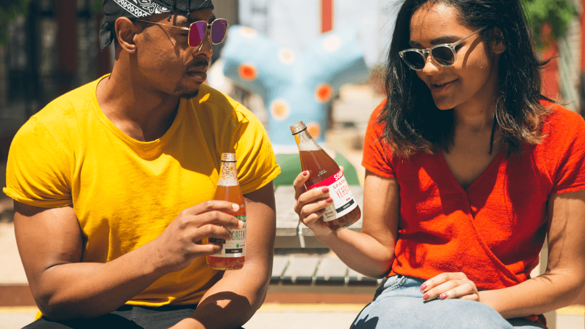 The idea of being bilingual represented by a Black man wearing a yellow t-shirt and a bandana along with a Latinx woman with long brown hair wearing a red shirt and sunglasses. They are talking to each other while sitting in a park and drinking some sort of tea out of glass bottles.