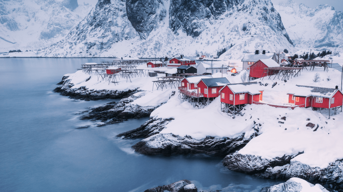 Winter survival secrets from Scandinavia represented by a photo of a small town in Scandinavia on the coast during winter. The town is filled with small red buildings on wooden stilts, and there are snowy mountains in the background.